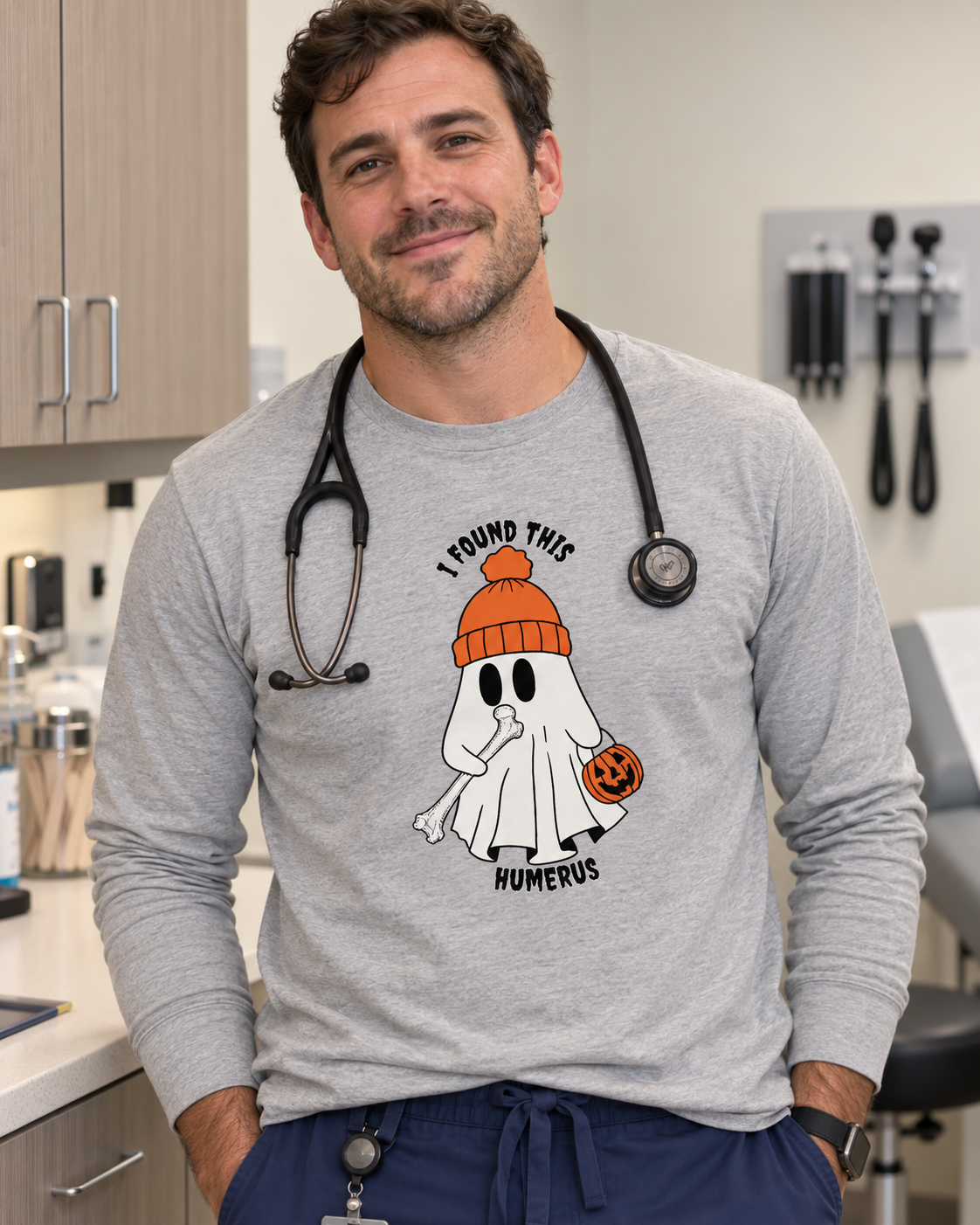 Male PA in heather grey long sleeve Halloween tee with ghost holding a humerus bone, wearing stethoscope in exam room