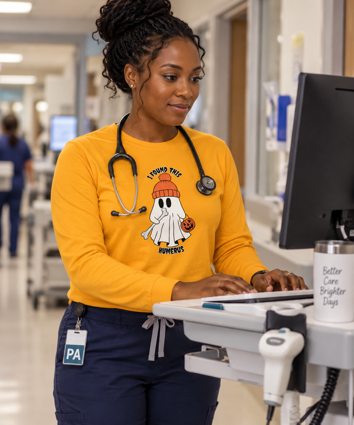 Female PA in gold long sleeve Halloween tee with ghost holding a humerus bone, wearing stethoscope at hospital workstation