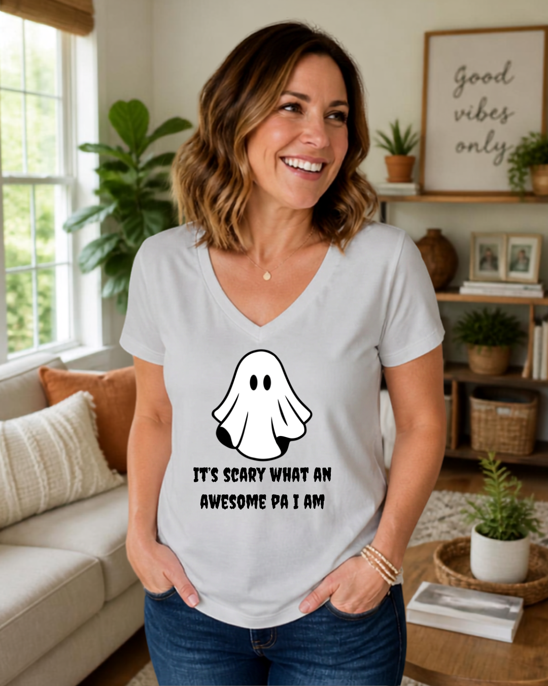 A woman wearing a white v-neck t-shirt with a black ghost graphic and the text 'IT'S SCARY WHAT AN AWESOME PA I AM' stands in a cozy living room setting.